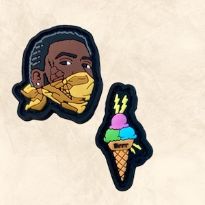 May include: Two black and yellow rubber patches. One patch features a cartoon portrait of a person wearing a yellow bandana. The other patch features a cartoon ice cream cone with three scoops of ice cream in pink, green, and blue colors. The ice cream cone has the word "Brrr" written on it.
