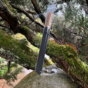 May include: A pack of incense sticks wrapped in clear plastic, resting on a mossy tree branch and a large rock. The incense is smoldering and producing smoke.