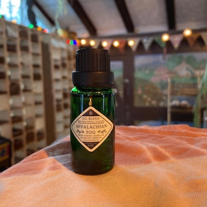 May include: A green glass essential oil bottle with a black cap. The label reads "Oil Blend Appalachian Fog" with text indicating ingredients and 1 oz. The bottle is on a peach-colored surface.