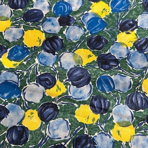 Op de afbeelding: Close-up van een stof met een herhalend bloemenpatroon. Het ontwerp toont blauwe, gele en marineblauwe bloemachtige vormen op een groene achtergrond. De stof heeft een getextureerde uitstraling, met de bloemen in een verspreide lay-out.