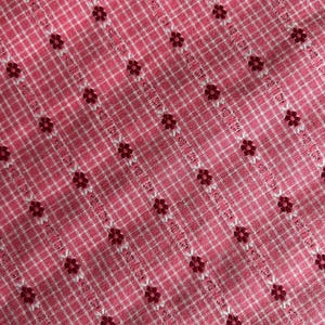 May include: Close-up of a pink and white checkered fabric with a floral pattern. The fabric has a grid-like pattern with small, dark red flowers evenly spaced throughout. The background is a light pink color.