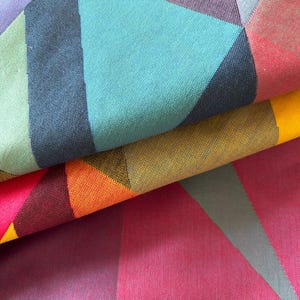 May include: Close-up of a folded fabric with a geometric pattern. The design features triangles in shades of teal, purple, pink, orange, yellow, and green. The fabric has a textured appearance, with visible woven threads.