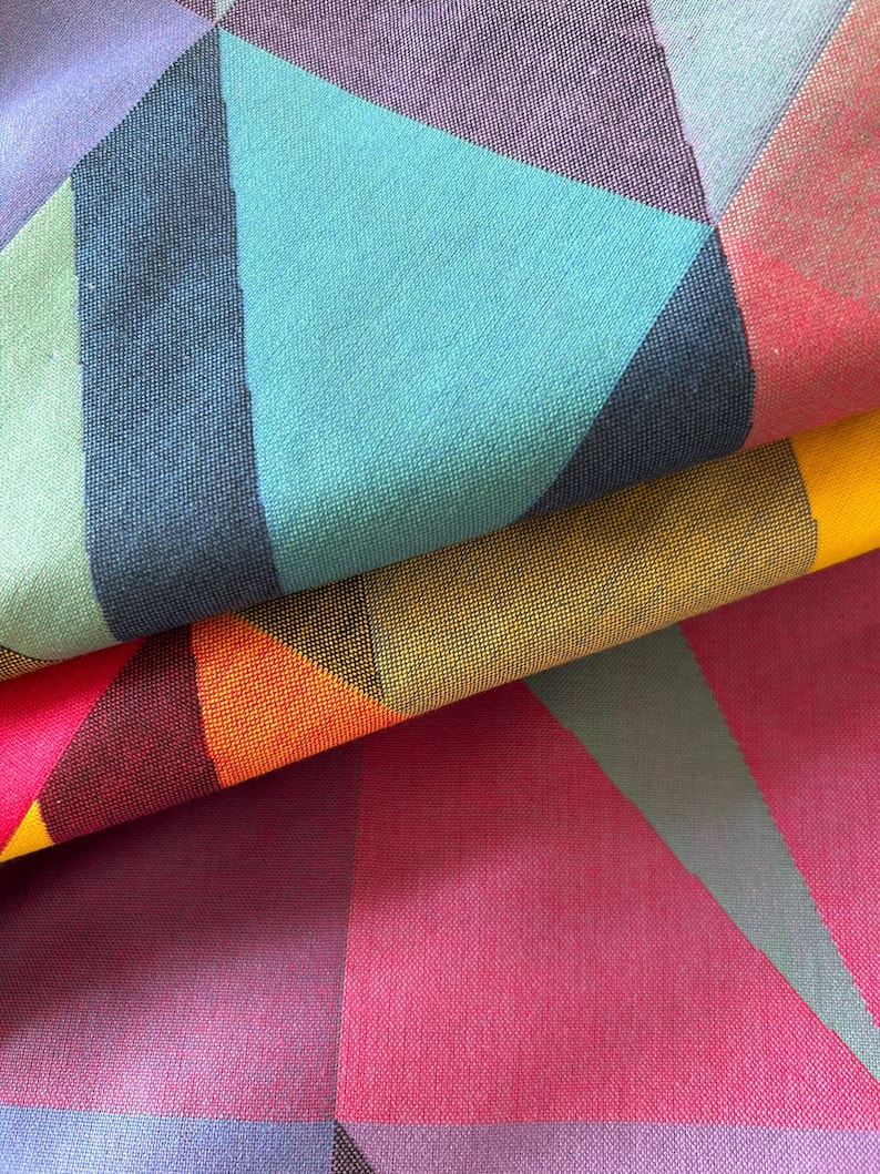 May include: Close-up of a folded fabric with a geometric pattern. The design features triangles in shades of teal, purple, pink, orange, yellow, and green. The fabric has a textured appearance, with visible woven threads.