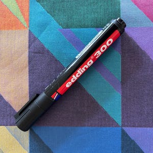 May include: A black Edding 300 permanent marker with a red band and white text, lying on a colorful geometric patterned fabric. The marker's cap is closed, and the brand name is prominently displayed.