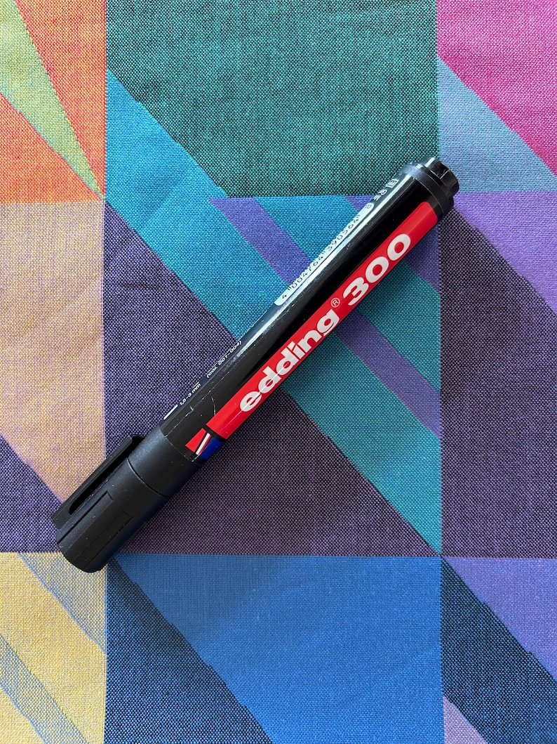 May include: A black Edding 300 permanent marker with a red band and white text, lying on a colorful geometric patterned fabric. The marker's cap is closed, and the brand name is prominently displayed.