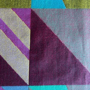 May include: Close-up of a textile with a geometric pattern. The design features large, angular shapes in shades of teal, purple, gray, and chartreuse. Diagonal stripes in purple and gray add further visual interest to the fabric.