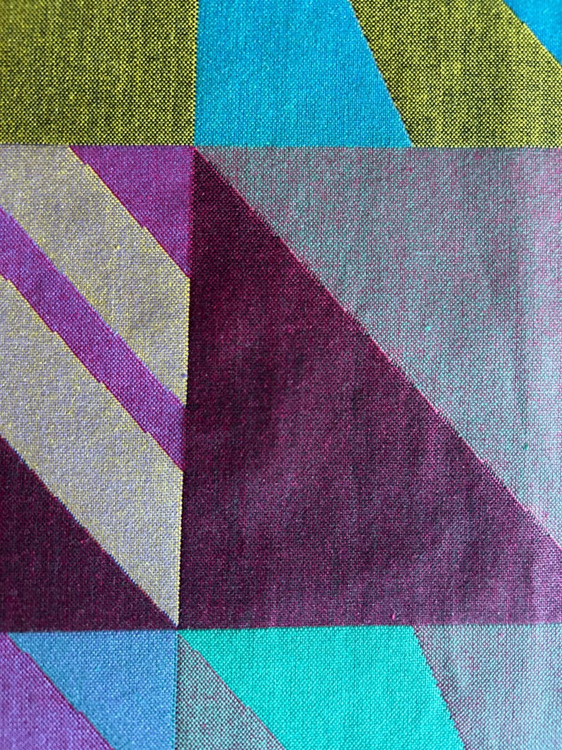 May include: Close-up of a textile with a geometric pattern. The design features large, angular shapes in shades of teal, purple, gray, and chartreuse. Diagonal stripes in purple and gray add further visual interest to the fabric.