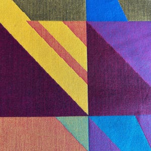 May include: A close-up of a textile with a geometric pattern. The design features squares and triangles in various colors, including yellow, red, blue, purple, and orange. The fabric has a woven texture.