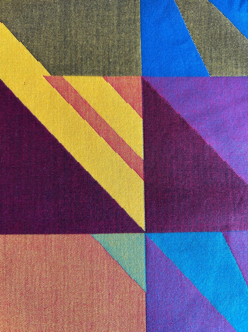 May include: A close-up of a textile with a geometric pattern. The design features squares and triangles in various colors, including yellow, red, blue, purple, and orange. The fabric has a woven texture.