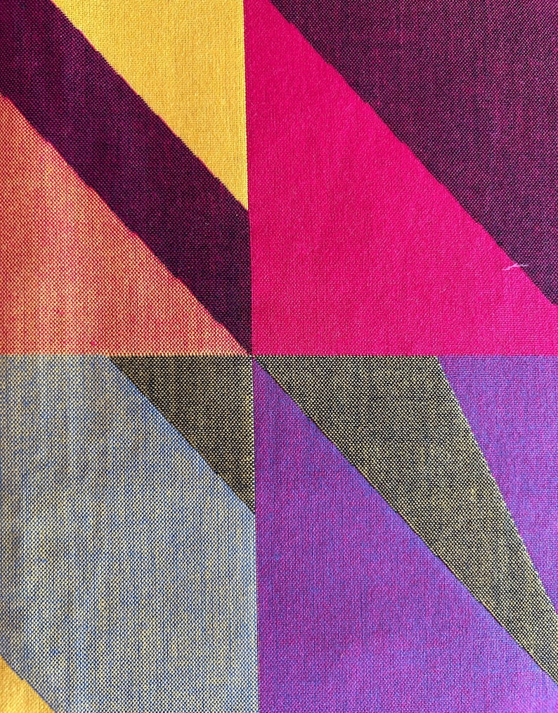 May include: Close-up of a geometric textile pattern. The design features large, angled shapes in vibrant colors, including yellow, orange, pink, purple, and gray. The fabric has a textured appearance, with visible woven threads.