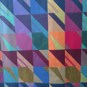 May include: A colorful textile with a geometric pattern. The design features squares and diagonal stripes in shades of blue, purple, green, orange, and pink. The fabric has a soft, slightly textured appearance.