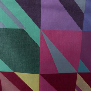 May include: Close-up of a fabric with a geometric pattern. The design features squares and triangles in shades of purple, pink, green, and yellow. The fabric has a textured appearance, suggesting a woven material. The overall aesthetic is modern and colorful.