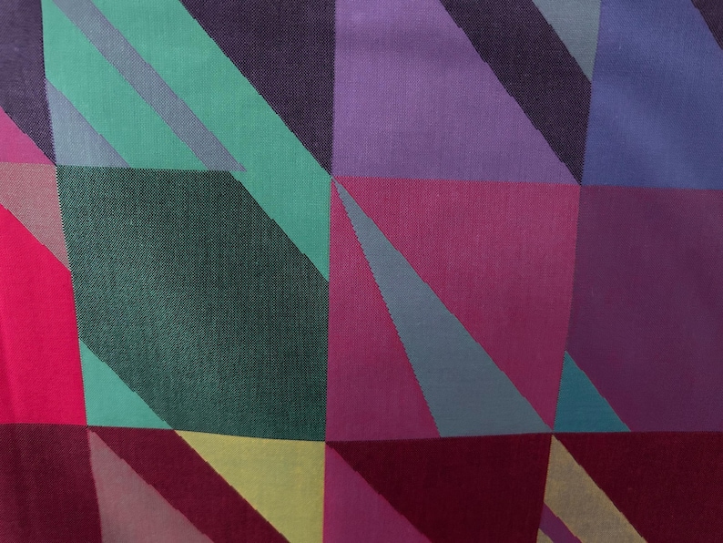 May include: Close-up of a fabric with a geometric pattern. The design features squares and triangles in shades of purple, pink, green, and yellow. The fabric has a textured appearance, suggesting a woven material. The overall aesthetic is modern and colorful.