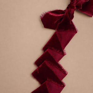 May include: A rich burgundy velvet ribbon with a frayed edge detail. The ribbon is tied into a bow at the top and falls in a zig-zag pattern. The background is a neutral beige colour.