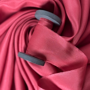 May include: A spool of red satin ribbon with grey plastic spools. The ribbon is lying on a folded piece of red satin fabric.