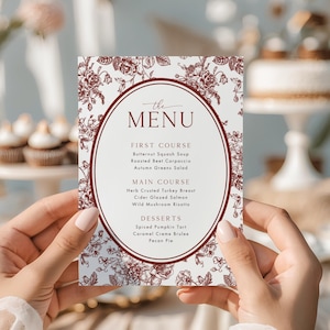 May include: A white menu card with a floral design and burgundy text. The menu lists a first course of butternut squash soup, roasted beet carpaccio, and autumn greens salad. The main course includes herb crusted turkey breast and cider glazed salmon.