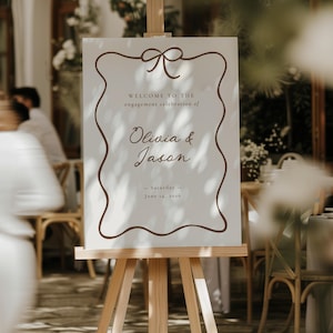 Op de afbeelding: Een wit bord op een houten ezel met bruine tekst en een decoratieve rand. Op het bord staat "Welkom bij de verlovingsviering van Olivia & Jason - Zaterdag - 14 juni 2016."