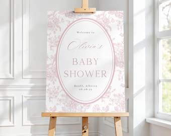 May include: A baby shower sign displayed on a wooden easel. The sign has a floral design in pale pink and white. The text reads "Welcome to Olivia's BABY SHOWER" with the location and date below. The easel is made of light-coloured wood.