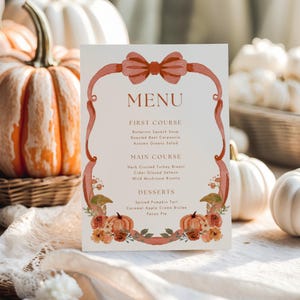 May include: A menu card with a watercolor design featuring a bow, ribbon, pumpkins, and floral accents. The menu lists courses such as Butternut Squash Soup, Herb Crusted Turkey Breast, and Spiced Pumpkin Tart. The card is set against a backdrop of pumpkins.