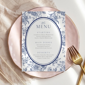 May include: A menu card with a floral design in blue and white, featuring an oval frame with the text "The Menu." The menu lists starters, main dishes, and desserts. The card is placed on a pink plate with a gold rim, accompanied by a gold fork.
