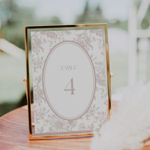 May include: A gold-framed table number card. The card features a floral pattern in shades of gray and a central oval with the word "TABLE" above the number "4". The card is displayed on a wooden surface.