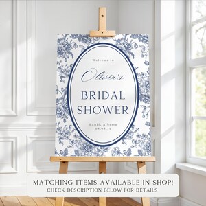 May include: A white sign with blue floral patterns and text that reads "Welcome to Olivia's Bridal Shower" with the date and location. The sign is displayed on a wooden easel. The text "Matching items available in shop! Check description below for details" is at the bottom.
