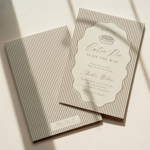 Puede incluir: Dos tarjetas de invitación rectangulares para baby shower con un patrón de rayas verticales en beige y blanco. La tarjeta frontal muestra un marco festoneado con una ilustración de pastel y el texto: A LITTLE CUTIE PIE IS ON THE WAY.