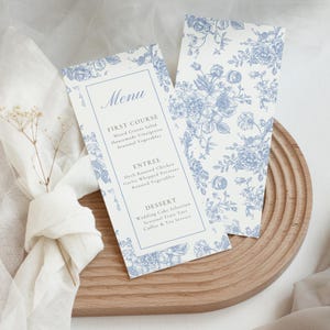 Peut inclure: Deux cartons de menu de mariage avec un motif floral bleu sur fond blanc. Le menu comprend les plats : Première entrée, Plat principal et Dessert. Les cartes sont sur un plateau en bois avec une serviette blanche et des fleurs séchées.