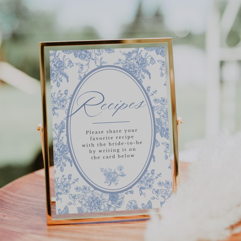 Pu&ograve; includere: Un cartello con cornice dorata con la parola "Ricette" in elegante corsivo. Il cartello presenta un design floreale blu e il testo: "Per favore, condividi la tua ricetta preferita con la futura sposa scrivendola sulla scheda qui sotto."