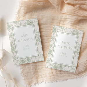 May include: Two wedding place cards with floral designs. Each card has a white rectangular frame with names and table information. The background features a light green floral pattern. The cards are on a beige fabric surface.