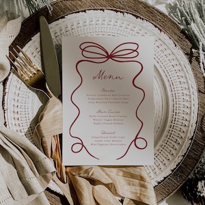 May include: A menu card with a burgundy bow design and text. The menu lists starters, main courses, and desserts. The card is on a white plate with a gold fork and knife. The plate is on a woven charger.