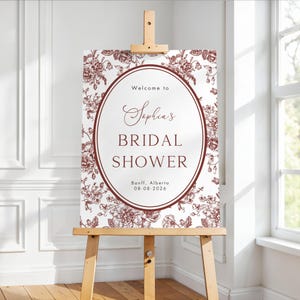 May include: A bridal shower sign displayed on a wooden easel. The sign has a burgundy and white floral design. The text reads "Welcome to Sophia's Bridal Shower", with the location and date beneath. The easel is made of light-coloured wood.
