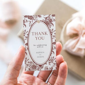 Può includere: Un'etichetta regalo rettangolare con un motivo floreale e le parole "THANK YOU" in un ovale centrale. L'etichetta include anche il testo "for celebrating with us" e i nomi "SOPHIA & JASON". L'etichetta ha un foro nella parte superiore.