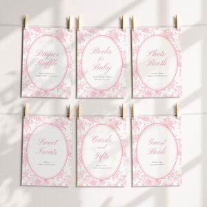 May include: Six pink floral-patterned signs with text for a baby shower, including "Diaper Raffle," "Books for Baby," "Photo Booth," "Sweet Treats," "Cards and Gifts," and "Guest Book." Each sign is held by a wooden clothespin.