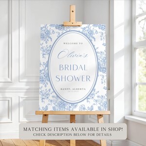 Pu&ograve; includere: Un cartello per l'addio al nubilato su un cavalletto di legno. Il cartello presenta un motivo floreale in blu chiaro e bianco, con il testo "Welcome to Olivia's Bridal Shower" e la data 15/08/2025. Banff, Alberta.