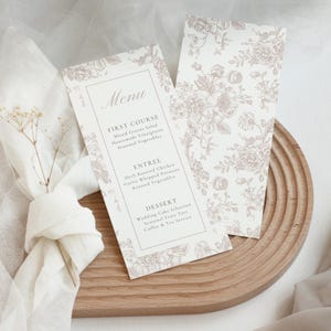 Puede incluir: Dos tarjetas de menú de boda con un diseño floral en un suave color malva. Las tarjetas de menú están sobre una tabla de servir de madera. El menú incluye los platos: Primer plato, Plato principal y Postre. El menú está impreso en una fuente serif clásica.