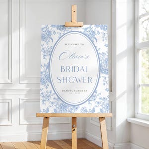 May include: A white bridal shower welcome sign with a blue floral pattern and a blue oval with the text "Welcome to Olivia's Bridal Shower, Banff, Alberta 08.13.2023".