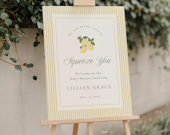 May include: A baby shower welcome sign on a wooden easel. The sign features a light yellow striped border and a cream-coloured centre with text and lemon illustrations. The text reads "Squeeze You" and welcomes guests to the baby shower honouring Lillian Grace.