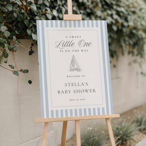 Puede incluir: Un letrero de bienvenida para un baby shower sobre un caballete de madera. El letrero tiene un borde a rayas azul claro y blanco. El texto dice "A Sweet Little One Is On The Way" con un gráfico de velero. Da la bienvenida a los invitados a Stella's Baby Shower, 14 de junio de 2026.