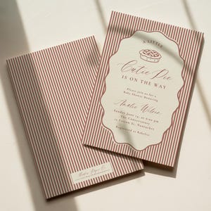 May include: Two rectangular baby shower invitation cards featuring a vertical red and white striped pattern. The front card has a scalloped frame containing a pie illustration and text reading A LITTLE CUTIE PIE IS ON THE WAY, honouring Amalie Wilson.