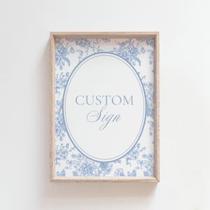 May include: A framed sign with a light wood frame. The sign features a white background with a blue floral pattern around the edges. An oval in the center contains the words "CUSTOM Sign" in blue script.