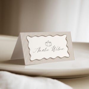 May include: A folded place card featuring a beige and white vertical striped pattern. The center has a scalloped rectangular frame containing a line drawing of a pumpkin and the printed name Amalie Wilson in a cursive script font.