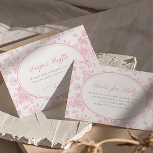Könnte beinhalten: Zwei Babyparty-Karten mit floralen Mustern. Auf einer Karte steht "Diaper Raffle" und auf der anderen "Books for Baby". Beide Karten haben rosa Blumendesigns auf weißem Hintergrund. Die Karten liegen auf einer strukturierten Oberfläche.
