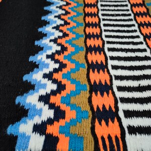 May include: Close-up of a colorful knitted fabric with geometric patterns in shades of blue, orange, white, and black.