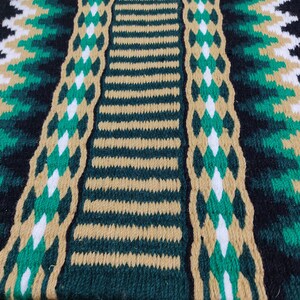 May include: Close-up of a woven saddle blanket with a black, green, and tan pattern. The blanket has a striped center with a diamond pattern on either side.