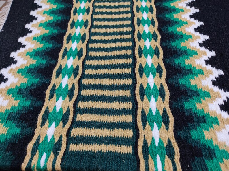 May include: Close-up of a woven saddle blanket with a black, green, and tan pattern. The blanket has a striped center with a diamond pattern on either side.