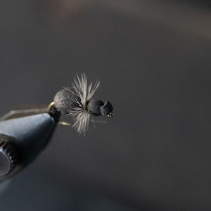 Puede incluir: Primer plano de un señuelo de pesca con mosca negro sujeto en un tornillo de banco. El señuelo tiene un cuerpo oscuro con alas gris claro y un pequeño anzuelo. El tornillo de banco es negro y plateado, sobre un fondo gris borroso.