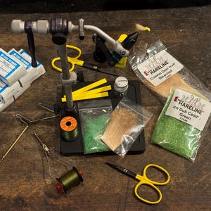 Könnte beinhalten: Eine Sammlung von Werkzeugen und Materialien zum Fliegenbinden, darunter ein Schraubstock, Faden, Schere und verschiedene Materialien. Das Bild zeigt Garnrollen, Materialbeutel und eine Auswahl an Werkzeugen, die alle auf einer Holzoberfläche angeordnet sind.