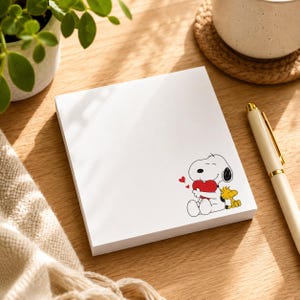 Puede incluir: Un bloc de notas cuadrado de color blanco con una ilustración impresa del personaje de dibujos animados Snoopy abrazando un corazón rojo mientras está sentado junto al pájaro amarillo Woodstock, con pequeños corazones rojos flotando arriba.