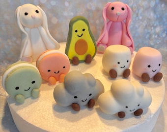 Op de afbeelding: Een verzameling grillige, kleurrijke fondant taarttoppers. De toppers omvatten een witte konijn, een roze konijn, een avocado, macarons, wolken en marshmallow-vormige figuren, allemaal met lachende gezichten. De toppers zijn gerangschikt op een witte taartbasis.